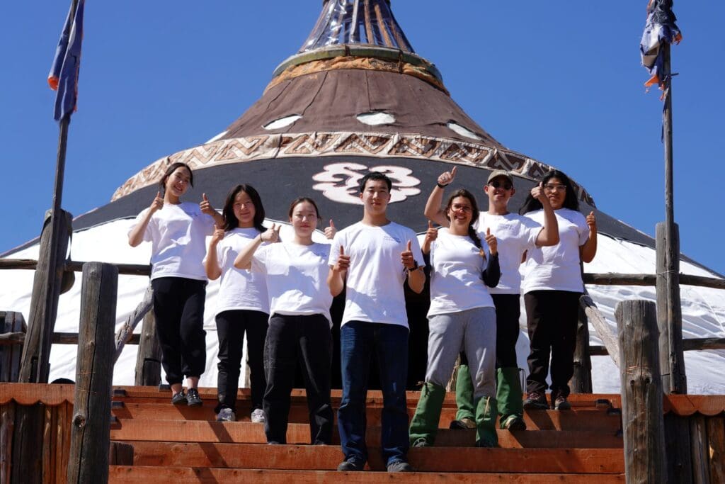 A group of seven young leaders wearing white t-shirts stand on wooden steps, giving thumbs up in front of a large tent-like structure in Mongolia under a clear blue sky, embodying empowerment and unity.