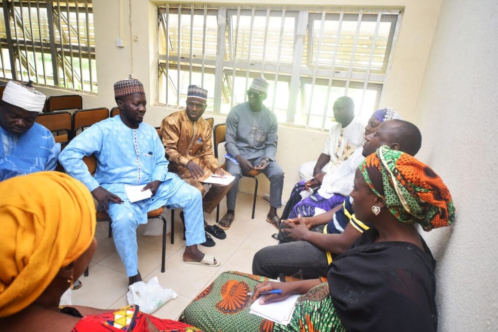 A group of people sit in a circle indoors, engaged in discussion about Interfaith Relationships, with notebooks and pens in hand, fostering community resilience as part of a GPF Nigeria initiative.