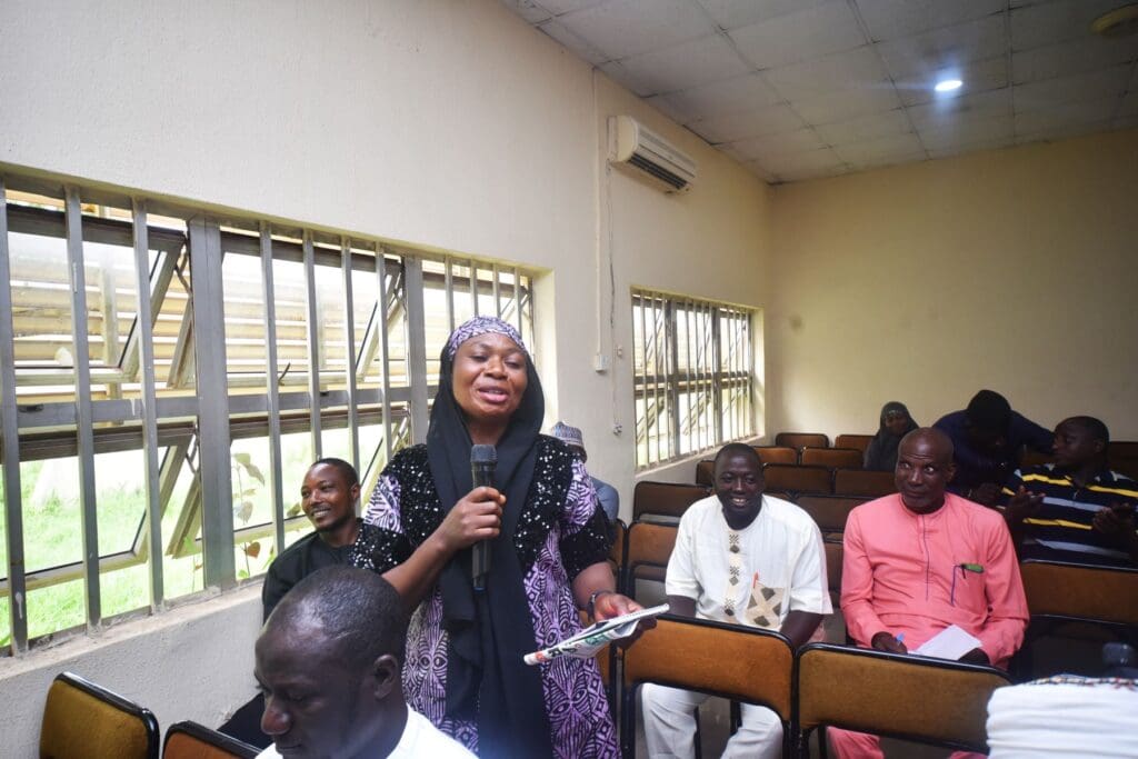 A woman in traditional attire speaks into a microphone while standing in a classroom with seated attendees, highlighting the importance of community resilience at a GPF Nigeria event.