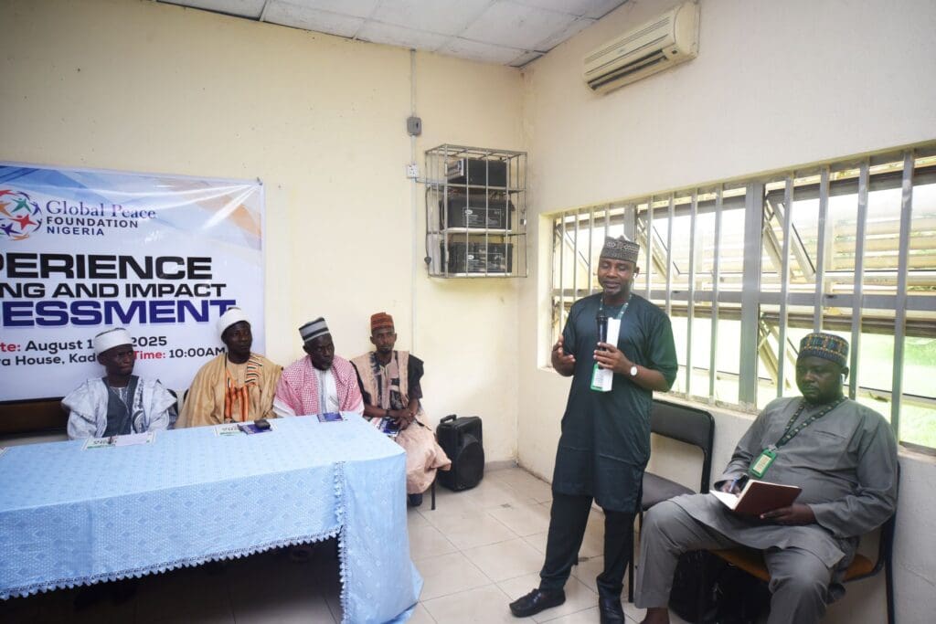 A group of men sit at a table while one man stands and speaks with a microphone during a GPF Nigeria event focused on “Experience Sharing and Impact Assessment,” highlighting community resilience.