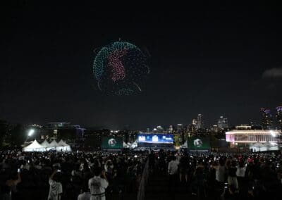 A nighttime outdoor event with a crowd watching a stunning drone show as lights form an image of the Korean Peninsula in the sky above the cityscape.
