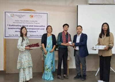 Five people stand in front of a banner at a graduation event for the Global Peacebuilders Leadership Program, exchanging gifts and certificates. The banner displays event details and logos of Global Peace Foundation and GPF Nepal.