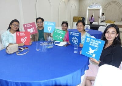 Five people sit around a table holding signs representing various United Nations Sustainable Development Goals in a conference room during the GPF Nepal Global Peacebuilders Leadership Program.