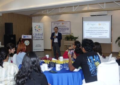 A man speaks on stage at a Global Peace Foundation event, such as the Global Peacebuilders Leadership Program, as attendees seated at round tables listen and take photos in a conference room.