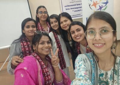 A group of seven young women, part of the Saksham Nari Fellows, pose for a selfie indoors, smiling at the camera, with a banner and whiteboard in the background.