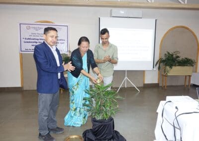 Three people are gathered indoors, clapping as a woman in a blue sari waters a potted plant during a formal Saksham Nari Fellows event by GPF Nepal.