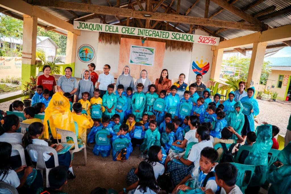 A group of school children from Camalaniugan and adults pose in a covered outdoor area, some kids in blue raincoats with backpacks. A school sign in the background hints at a partnership with GPF Philippines or St. Paul University Philippines.