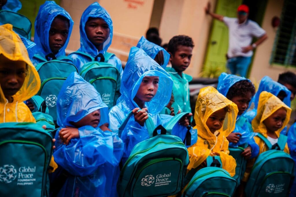 School children from Camalaniugan wearing blue and yellow raincoats hold green backpacks labeled 