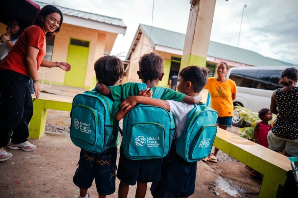 Three school children from Camalaniugan, wearing matching teal backpacks, stand arm-in-arm outside a building, with adults and other children nearby—an initiative supported by GPF Philippines and St. Paul University Philippines.