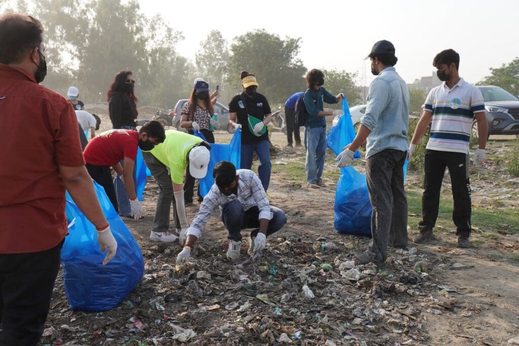 A group of people wearing gloves and masks collect trash and litter into blue bags during a GPF India community cleanup outdoors, supporting Yamuna river revival efforts.