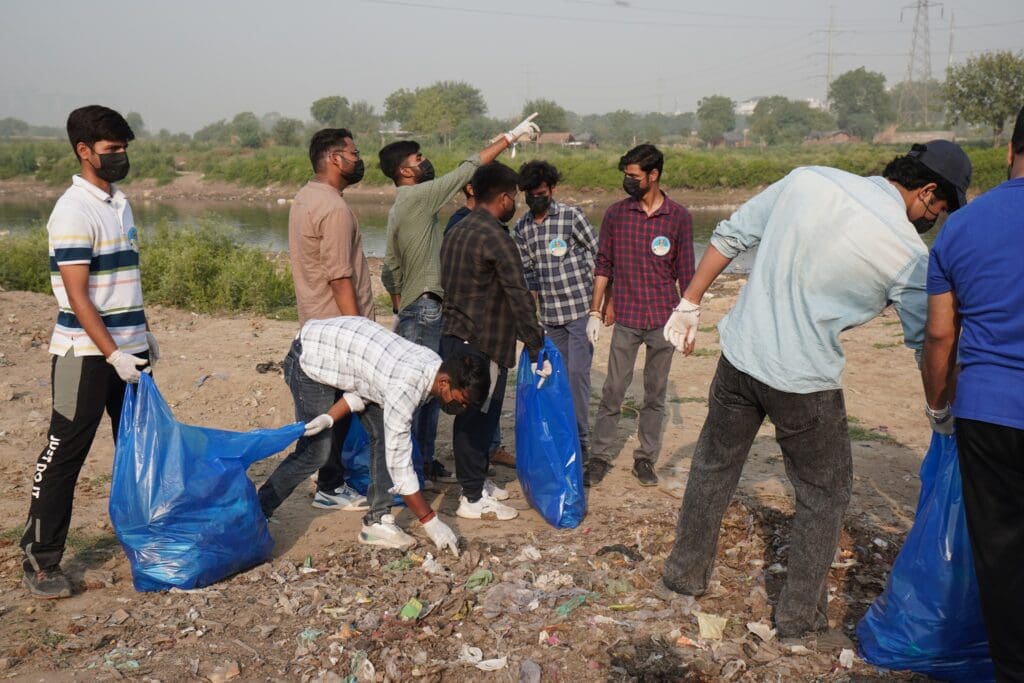 A group of people wearing gloves and masks collect trash and debris in blue bags during a River Revival cleanup by GPF India near the Yamuna riverbank.