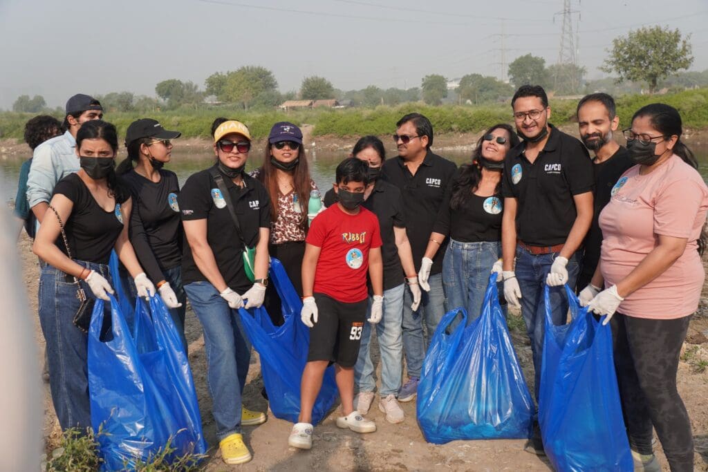 A group of people wearing gloves and holding large blue bags poses together outdoors near the Yamuna riverbank during a GPF India cleanup activity, supporting community efforts toward river revival.