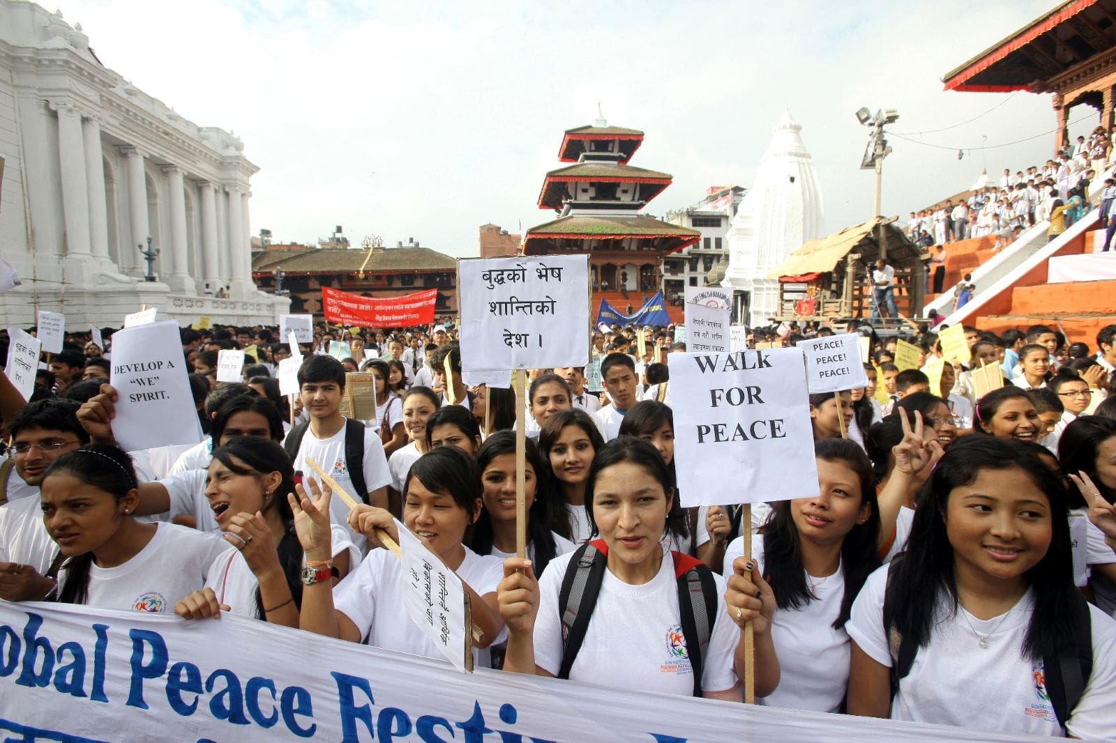 A large group of people in white shirts hold signs promoting peace during a public march for the International Day of Peace in an outdoor urban area with historic buildings.
