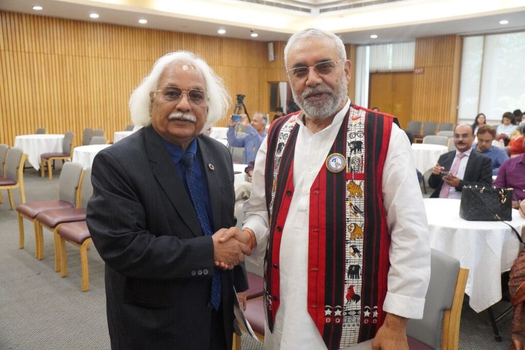 Two men shake hands in a conference room; one in a suit and tie, the other in traditional attire with a patterned vest. The scene highlights the importance of Youth Skills for building a Sustainable Future. Other people are seated in the background.