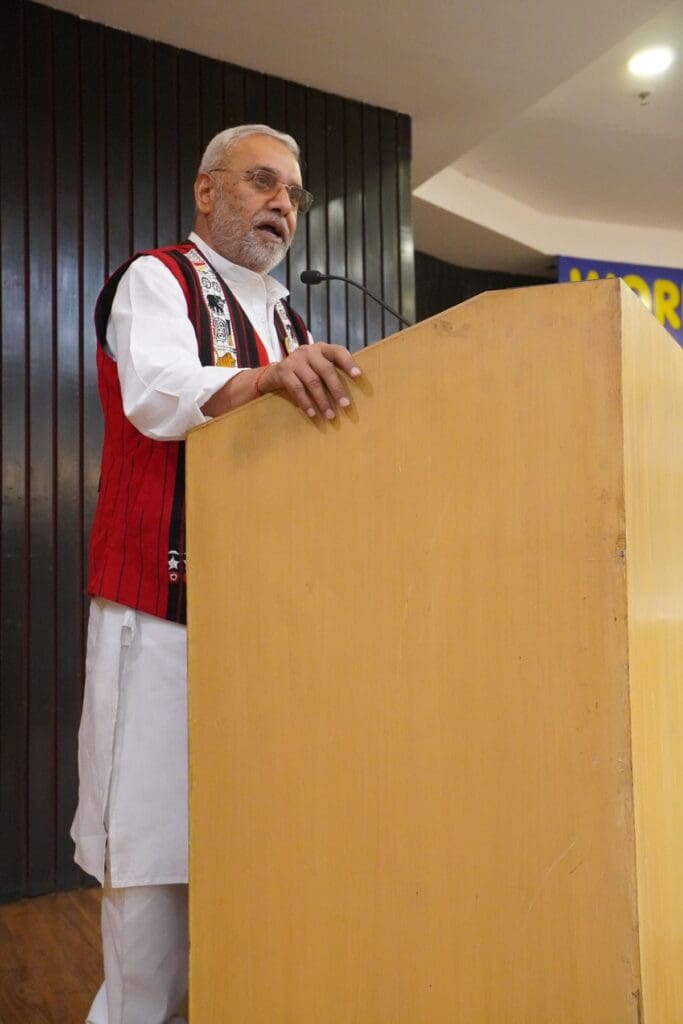 An older man wearing glasses and traditional attire speaks at a wooden podium with a microphone in an indoor setting, addressing the importance of Youth Skills for a Sustainable Future on World Youth Skills Day.