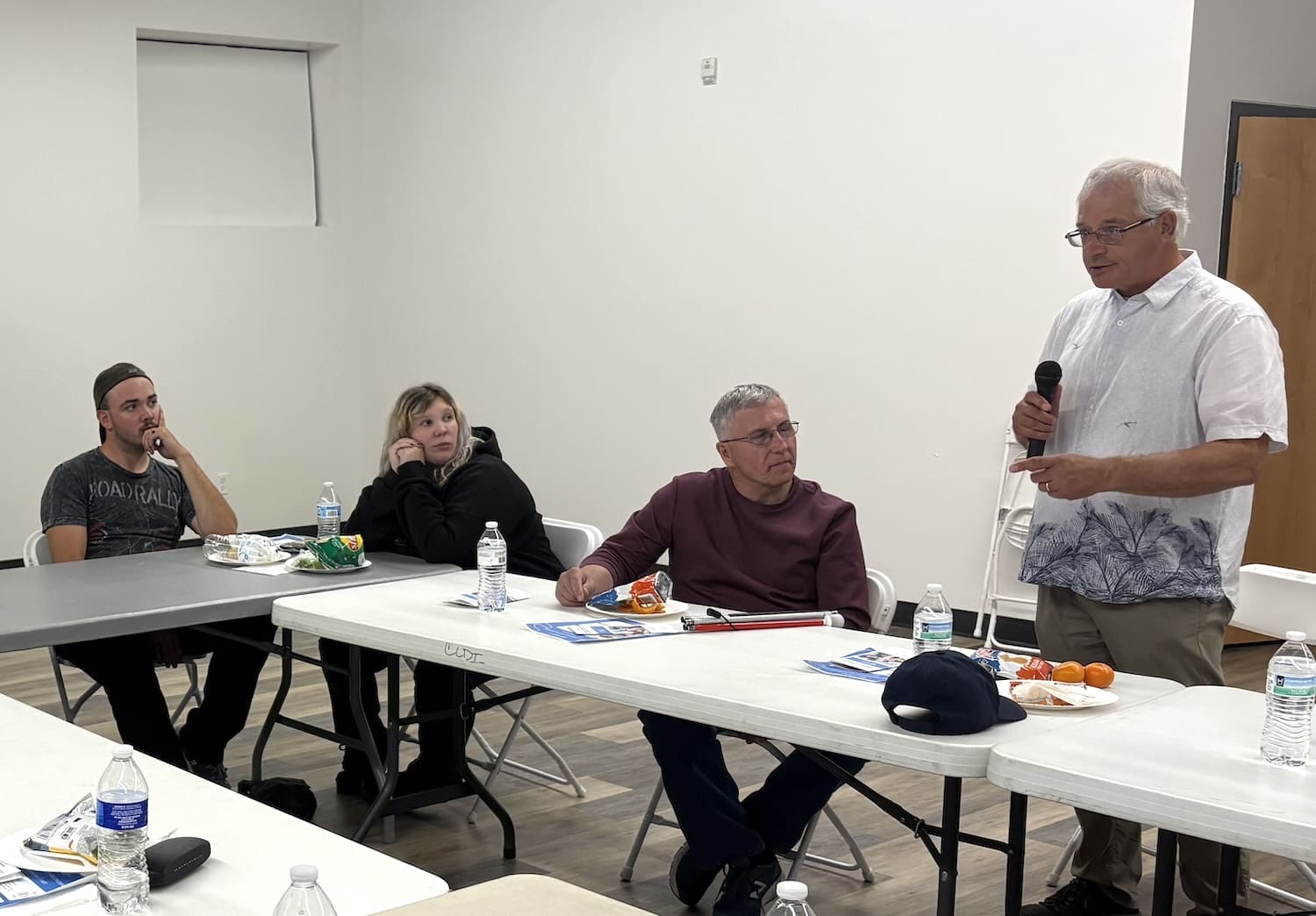 Michael Yakawich stands and speaks into a microphone in a plain Montana meeting room as three people seated at tables listen. Papers, water bottles, and snacks are on the tables during this discussion on community reconciliation.