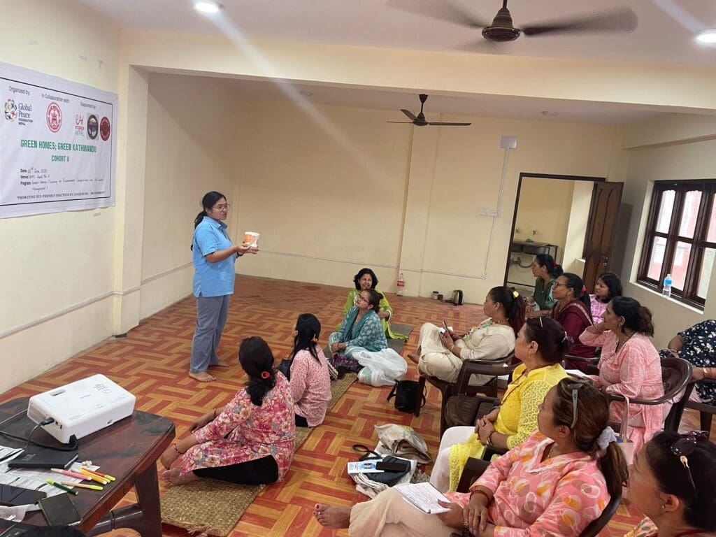 A woman stands and presents Eco-Friendly Training to a seated group of women in a classroom, as part of the Green Kathmandu Project, with a banner on the wall and a projector on the floor.