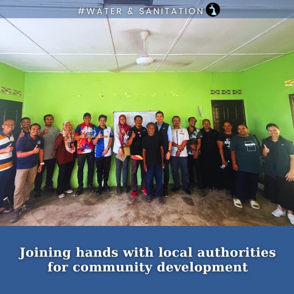 A group of people stands together indoors in front of a green wall, posing for a photo under a banner about clean water and sanitation, highlighting efforts to improve communities in Malaysia.