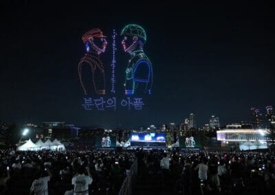 A nighttime crowd watches a mesmerizing drone show, featuring two soldiers facing each other with Korean text beneath the display, set against a vibrant city skyline backdrop.
