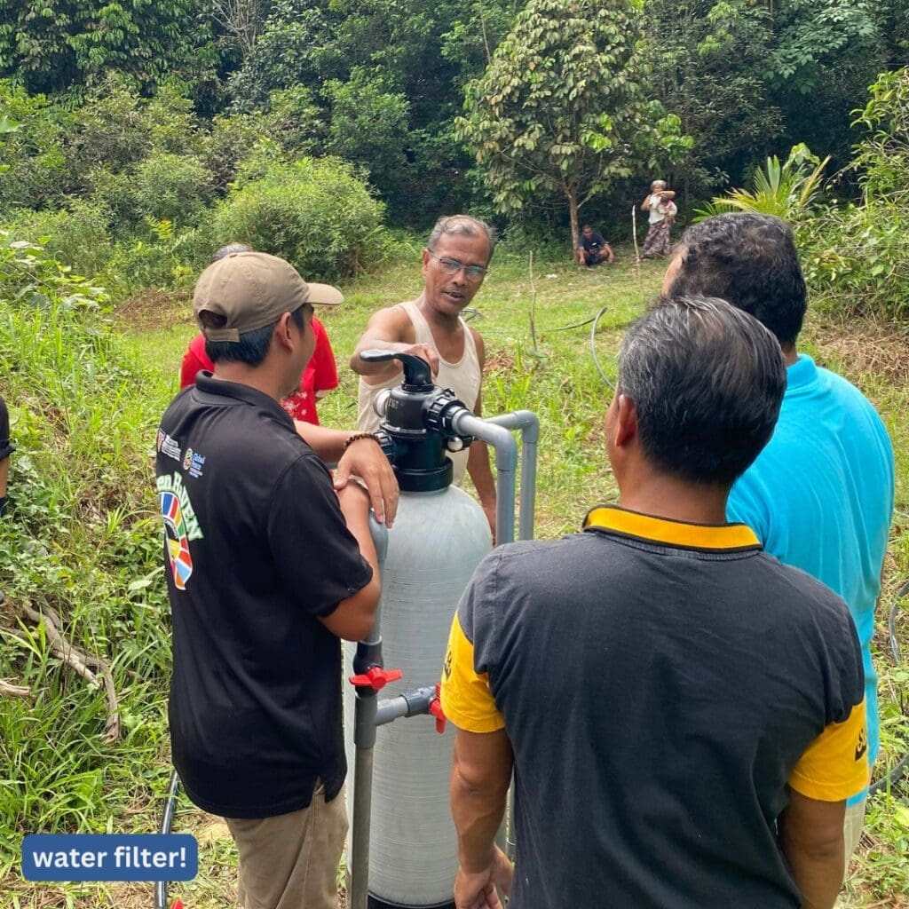 Several people stand around a water filtration system outdoors in a grassy, wooded area in Malaysia, while one person explains how it improves community access to clean water.