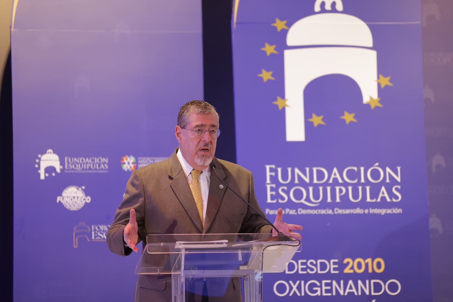 A man in a suit speaks at a clear podium in front of a blue Fundación Esquipulas banner, featuring white and yellow text and a stylized arch, during the Esquipulas Regional Forum on democracy and regional integration.