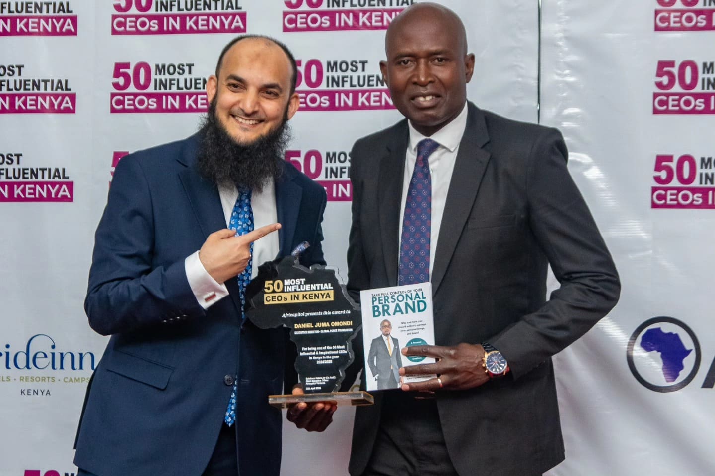 Two men in suits pose for a photo; Daniel Juma, Africa Regional Representative, holds an award plaque while the other holds a book titled 