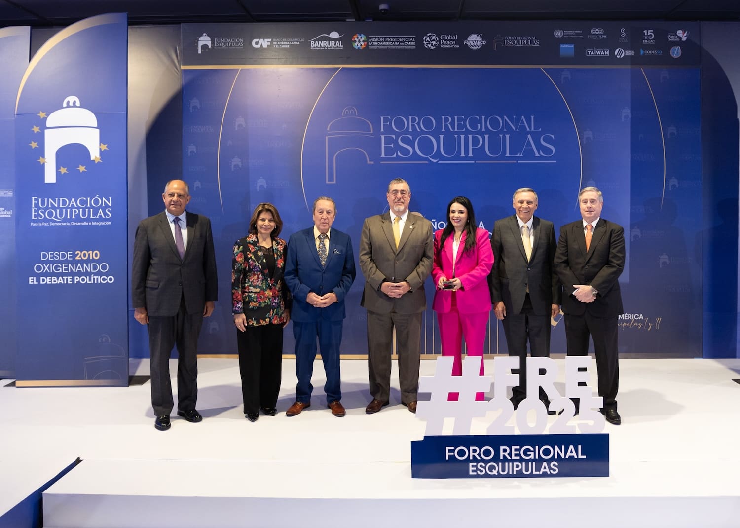 Seven people in formal attire stand in a row at the Esquipulas Regional Forum 2025, in front of a blue backdrop with event logos and a large hashtag sign, highlighting regional integration and strengthening democracy.