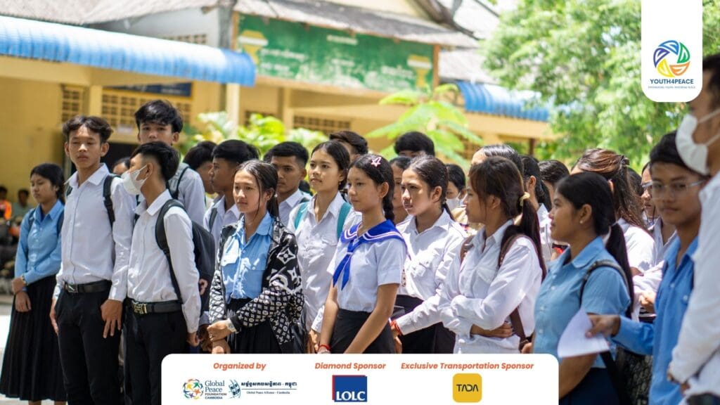 A group of Cambodian Youth in uniforms stand outdoors, some holding papers, with school buildings and trees in the background. Event sponsor logos are visible at the bottom, highlighting their participation in a Youth Development Program.