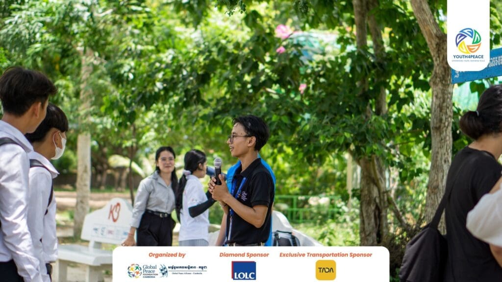 A man speaks into a microphone to a small group outdoors, with trees and event sponsor logos visible, inspiring leadership and growth among Cambodian youth.