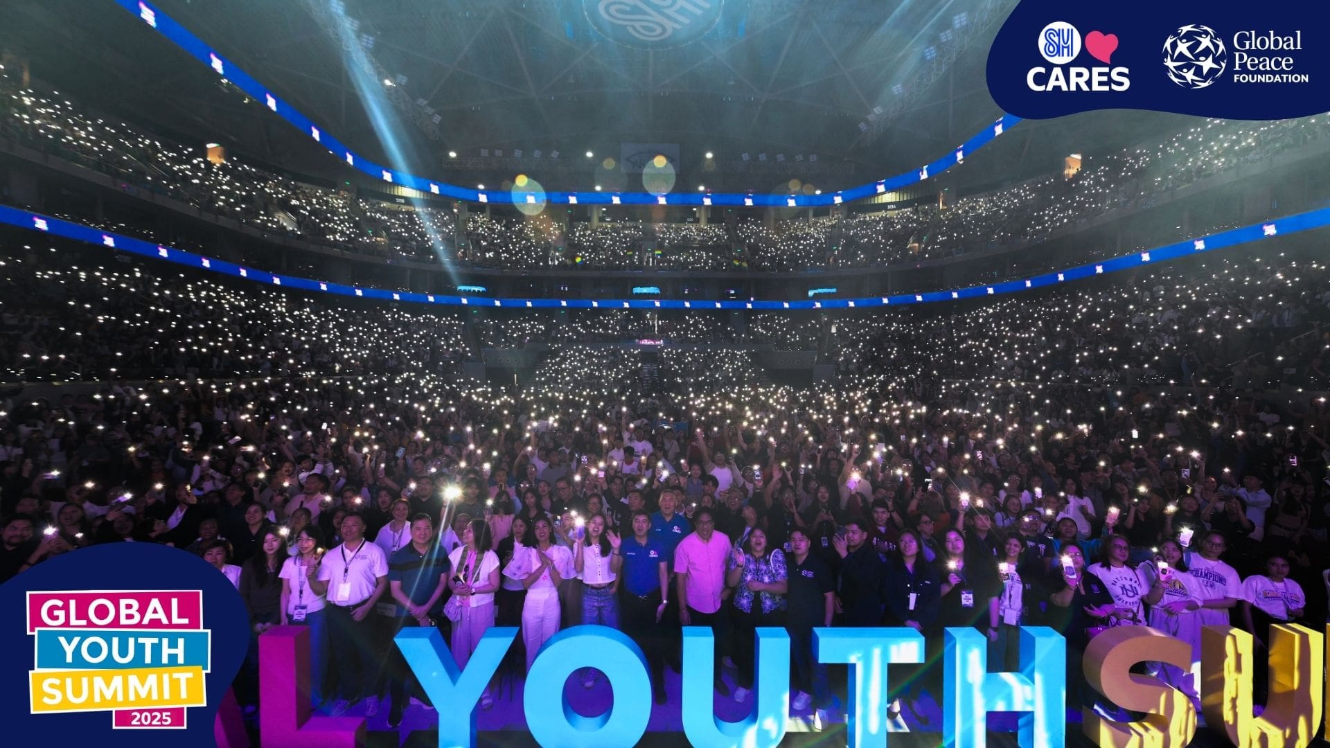 A large crowd holds up phone flashlights in a brightly lit stadium at the Global Youth Summit 2025, where Young Filipinos unite for Positive Change, with event logos and 