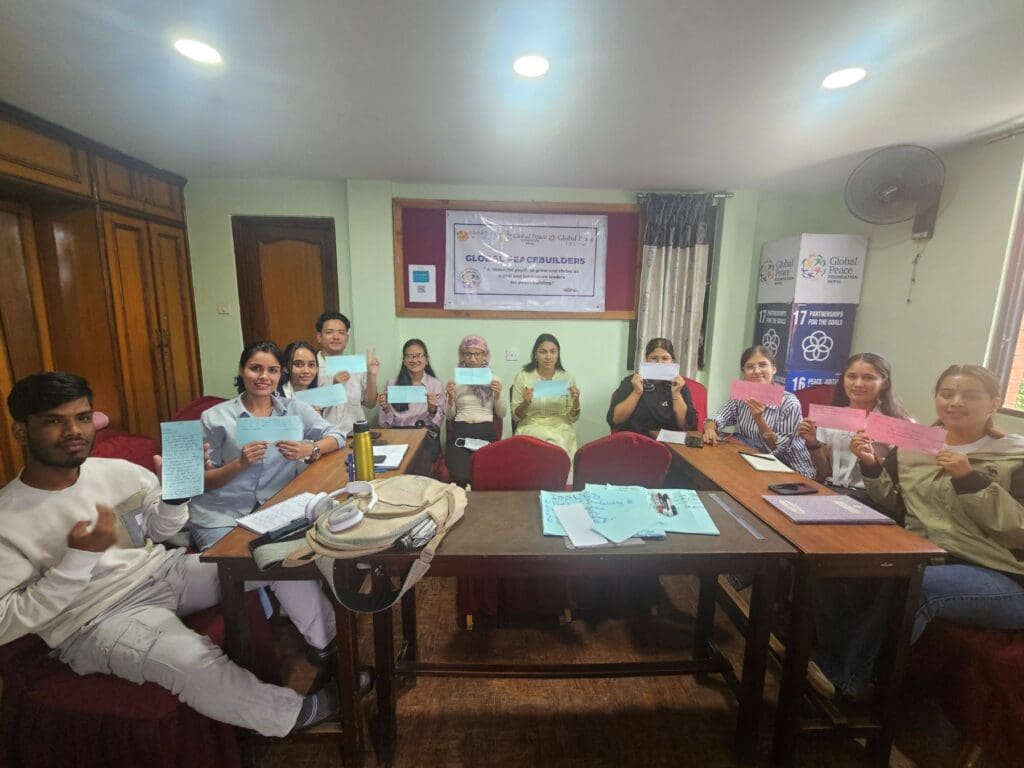 A group of people sit around a conference table holding up papers, with informational posters and a Nepal International Day of Peace 2025 banner on the wall behind them.
