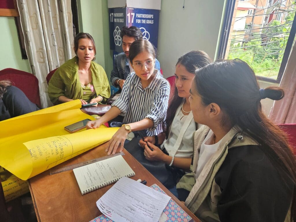 Four young women sit around a table discussing notes and writing on large yellow paper sheets during a group activity indoors for Nepal International Day of Peace 2025.
