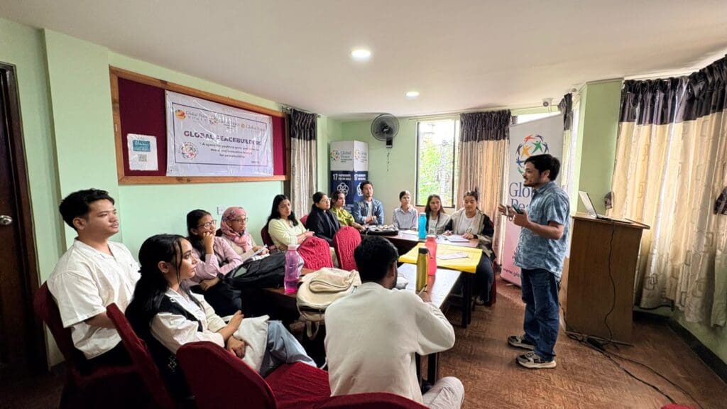 A group of people sit around a table listening to a man standing and speaking in a brightly lit room decorated with banners and curtains during the Nepal International Day of Peace 2025.