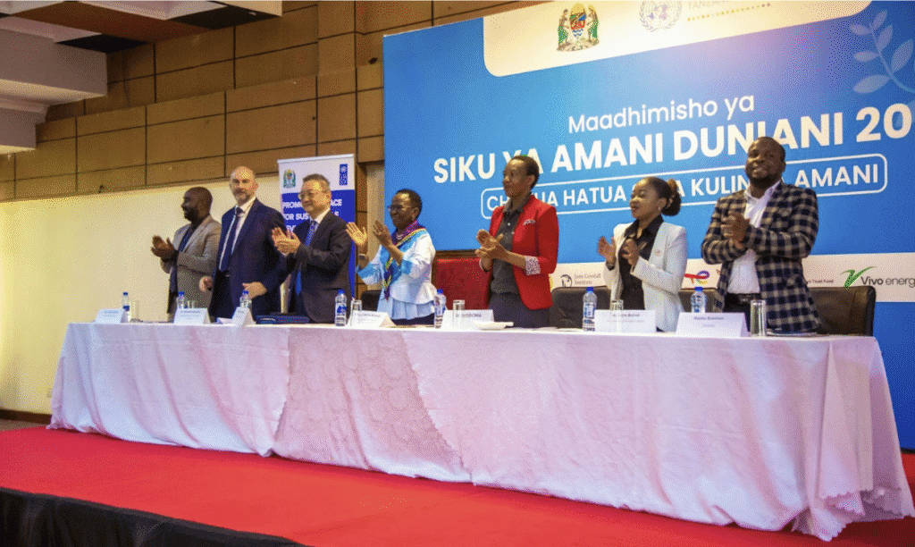 Seven people stand and applaud behind a table at a formal event in Tanzania, with a blue banner in Swahili about International Peace Day and building a Peaceful World displayed in the background.