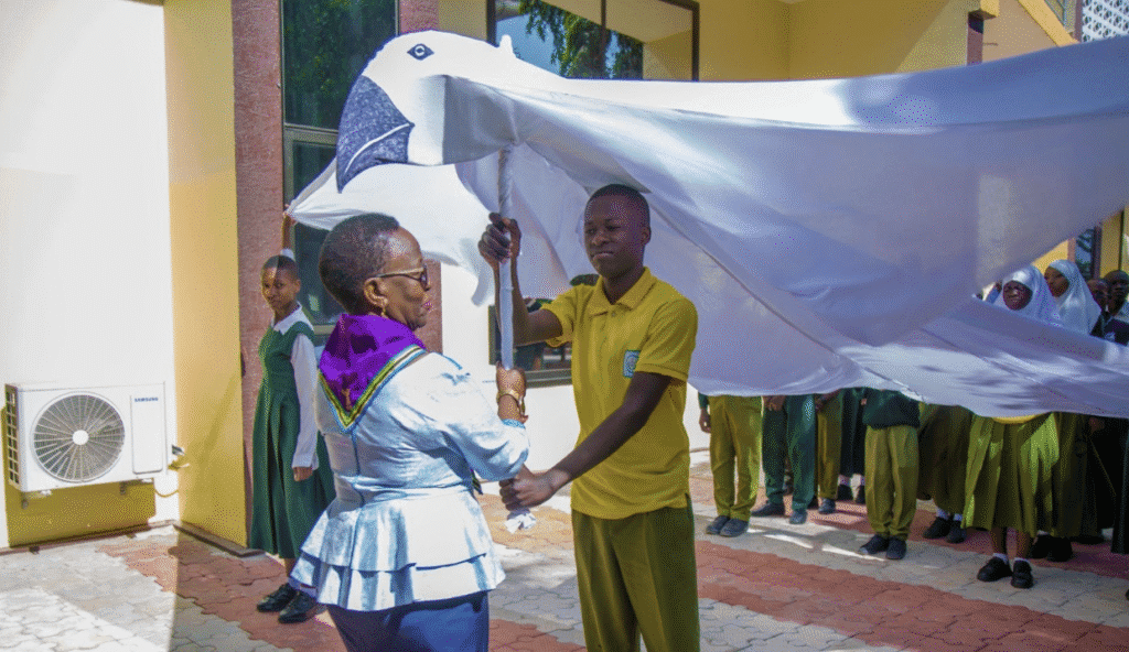 A woman and a student hold up a large white dove prop outdoors in Tanzania, with other students in uniform standing in the background to mark International Peace Day and promote a peaceful world.