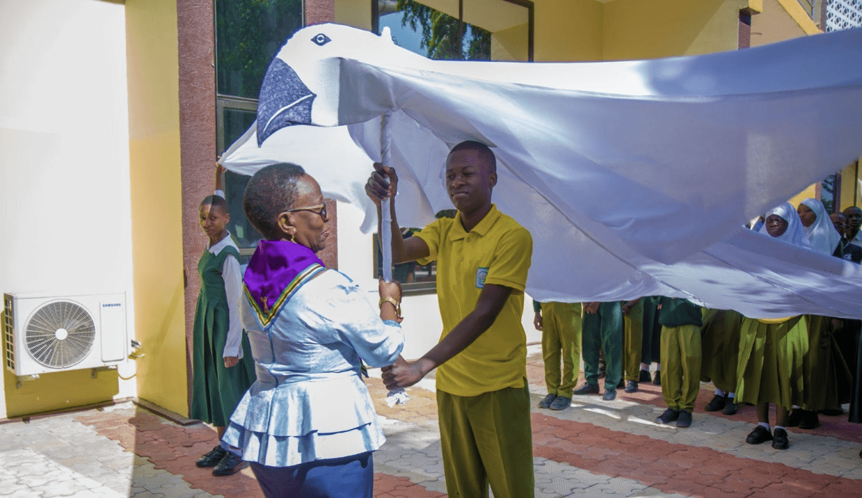 A woman and a student hold up a large white dove prop outdoors in Tanzania, with other students in uniform standing in the background to mark International Peace Day and promote a peaceful world.