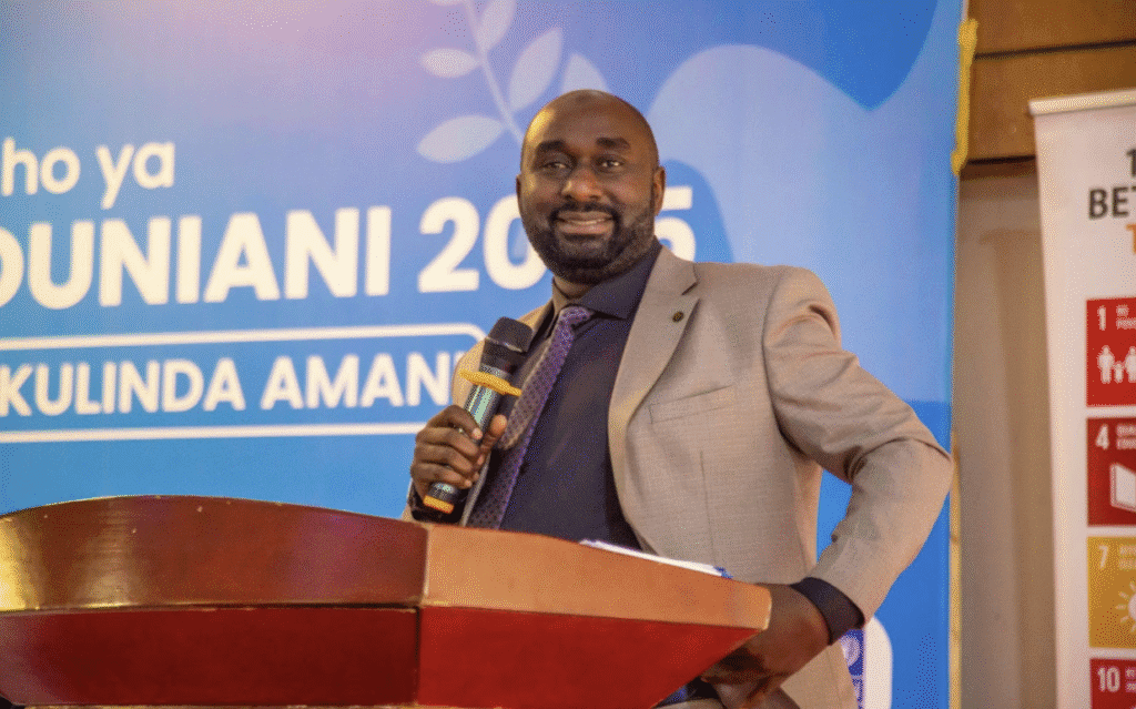 A man in a suit stands at a podium holding a microphone, smiling, with a blue event banner and a Sustainable Development Goals poster in the background—celebrating International Peace Day and the vision of a Peaceful World in Tanzania.
