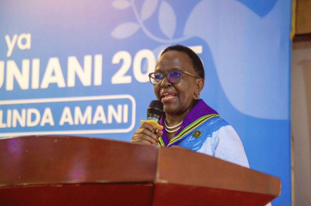 A woman in glasses and a colorful scarf speaks into a microphone at a podium in front of a blue International Peace Day event banner, sharing her hopes for a peaceful world in Tanzania.