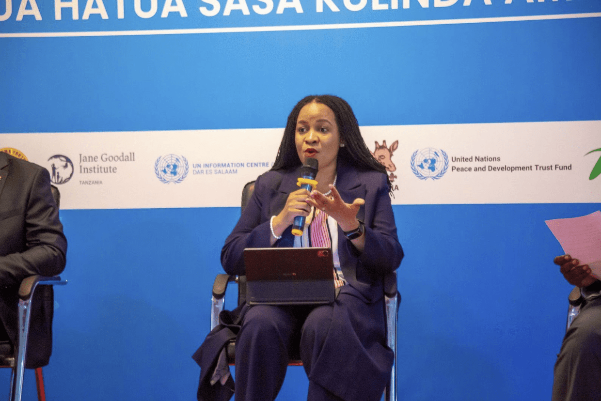 A woman in a suit speaks into a microphone while seated on a panel, with a blue backdrop displaying various organization logos behind her, discussing initiatives for a Peaceful World on International Peace Day in Tanzania.