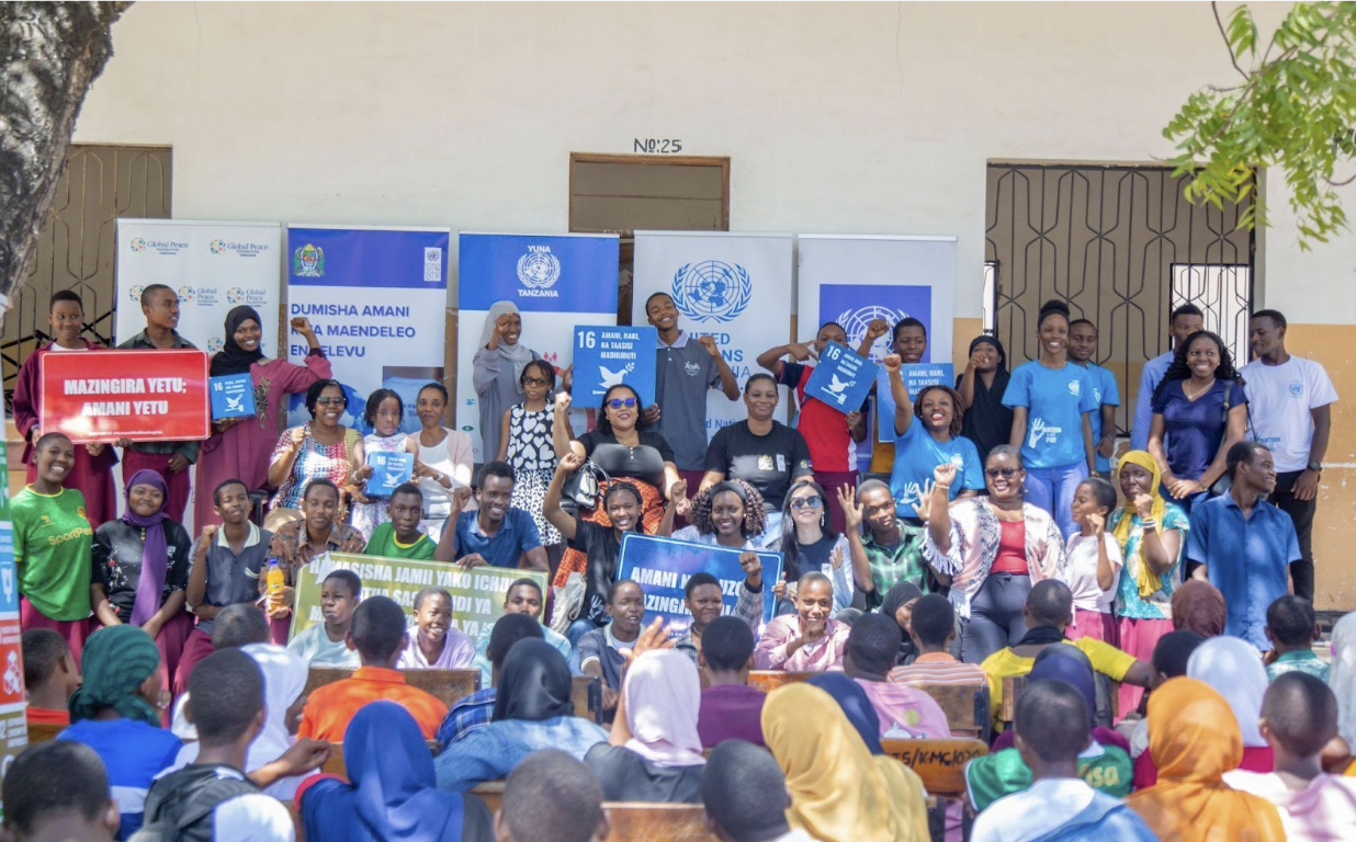 A large group of people pose for a photo in front of banners displaying United Nations logos and messages about peace and development in Tanzania, while an audience sits facing them, celebrating International Peace Day.