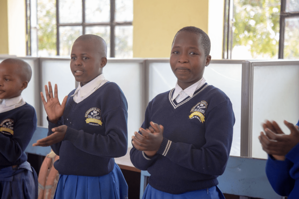 Four students in blue and white school uniforms stand indoors, clapping their hands in celebration of International Peace Day. The classroom, located in Tanzania, features large windows and light yellow walls.