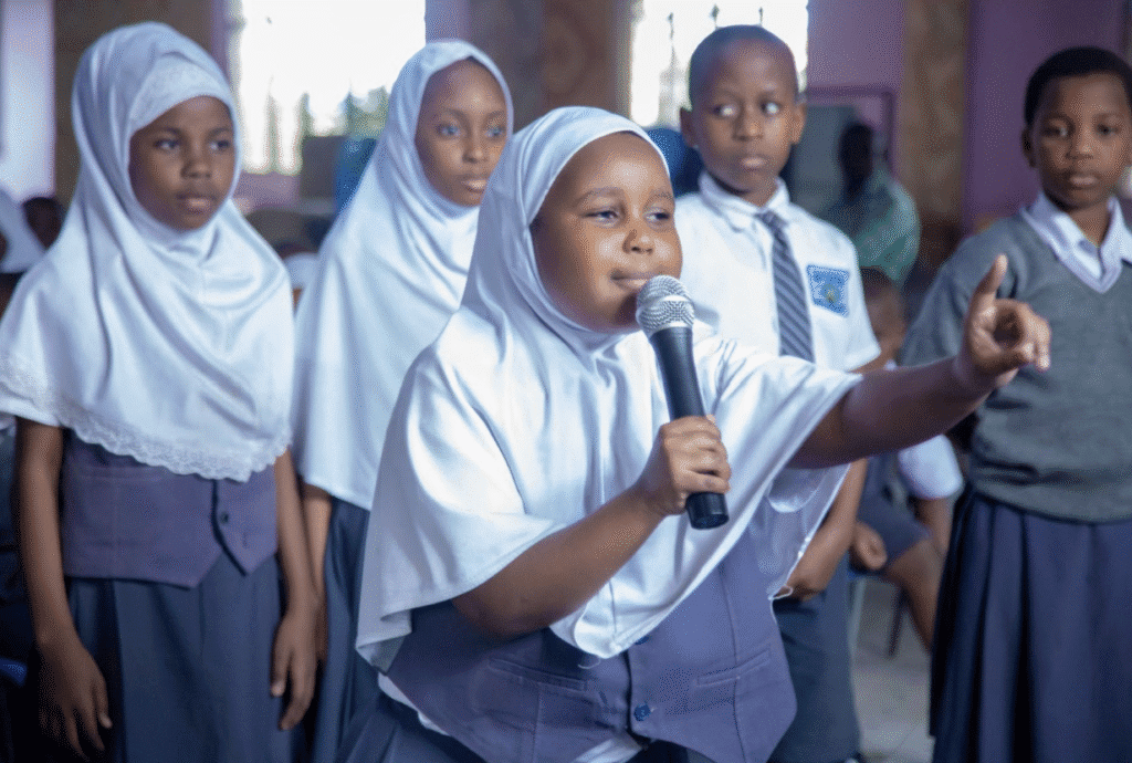 A young girl in a white hijab speaks into a microphone while other schoolchildren stand nearby in a classroom setting, celebrating International Peace Day in Tanzania.