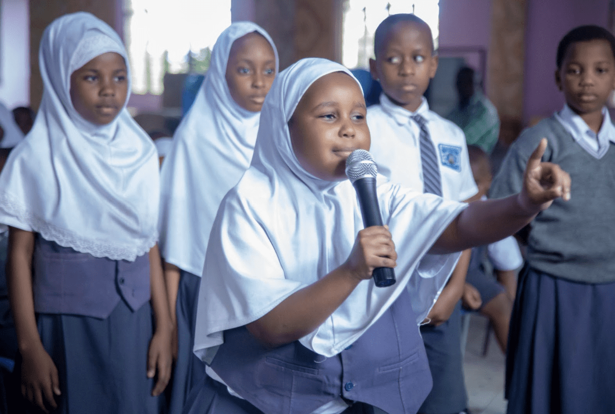A young girl in a white hijab speaks into a microphone while other schoolchildren stand nearby in a classroom setting, celebrating International Peace Day in Tanzania.
