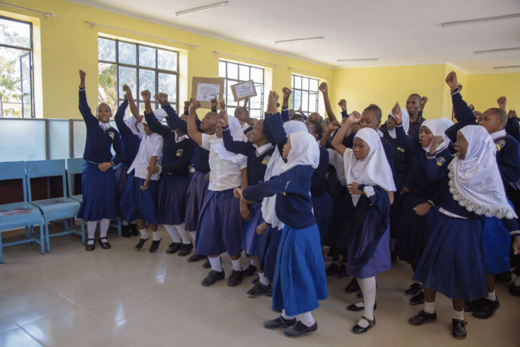 A group of students in school uniforms raise their fists and cheer inside a bright classroom with yellow walls and large windows, celebrating International Peace Day and dreaming of a more peaceful world.