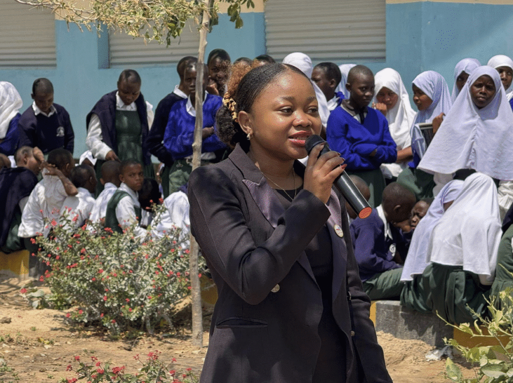 A woman in a black blazer speaks into a microphone outdoors in Tanzania, with a group of students in uniforms and hijabs seated behind her, marking International Peace Day.