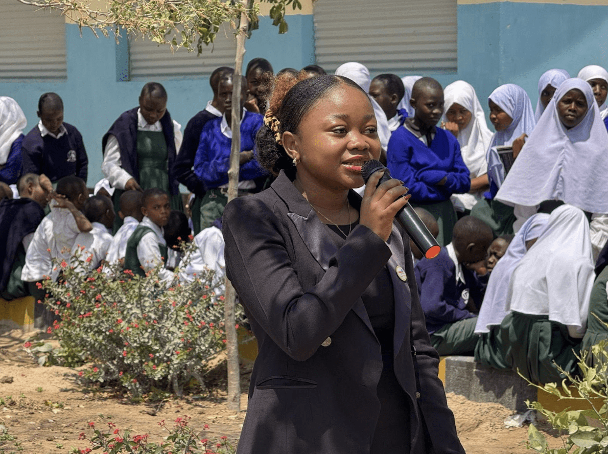 A woman in a black blazer speaks into a microphone outdoors in Tanzania, with a group of students in uniforms and hijabs seated behind her, marking International Peace Day.