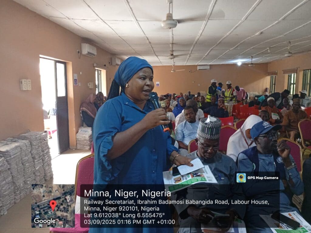 A woman speaks into a microphone at an indoor peacebuilding event in Minna, Niger State, with seated attendees listening. A location and timestamp overlay is visible in the image.