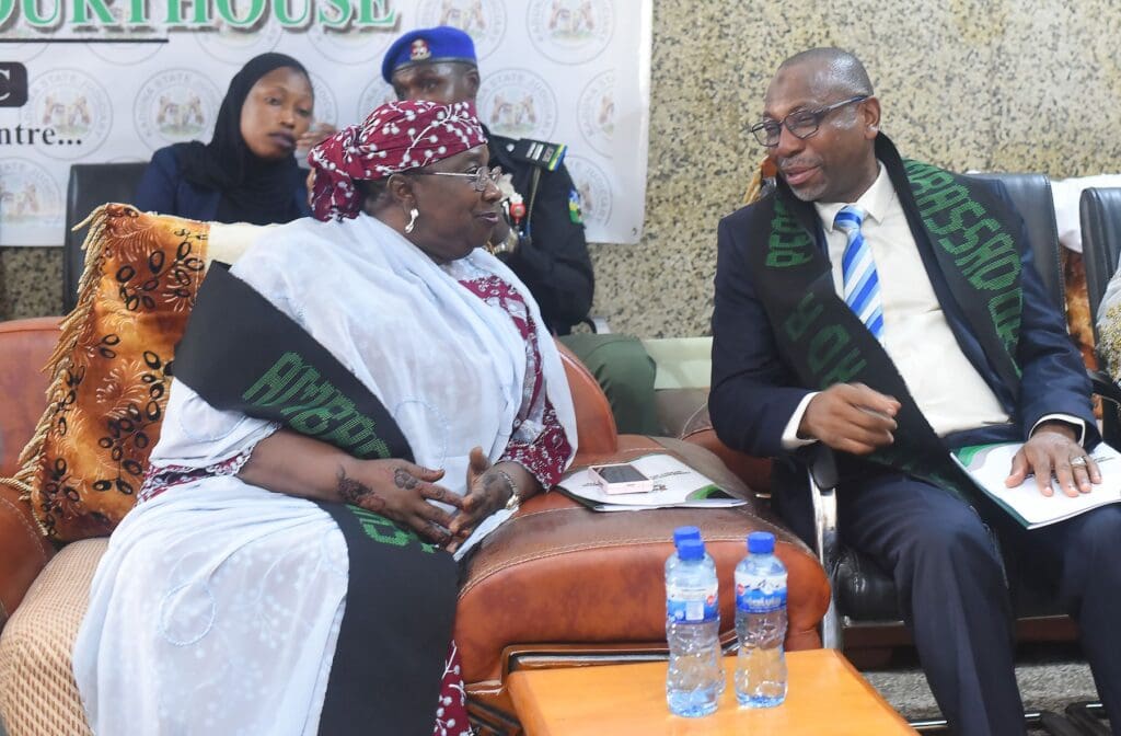 A woman and a man wearing black sashes sit and talk on a couch at an indoor event in Kaduna, discussing Alternative Dispute Resolution, with two water bottles on the table in front of them and others in the background.