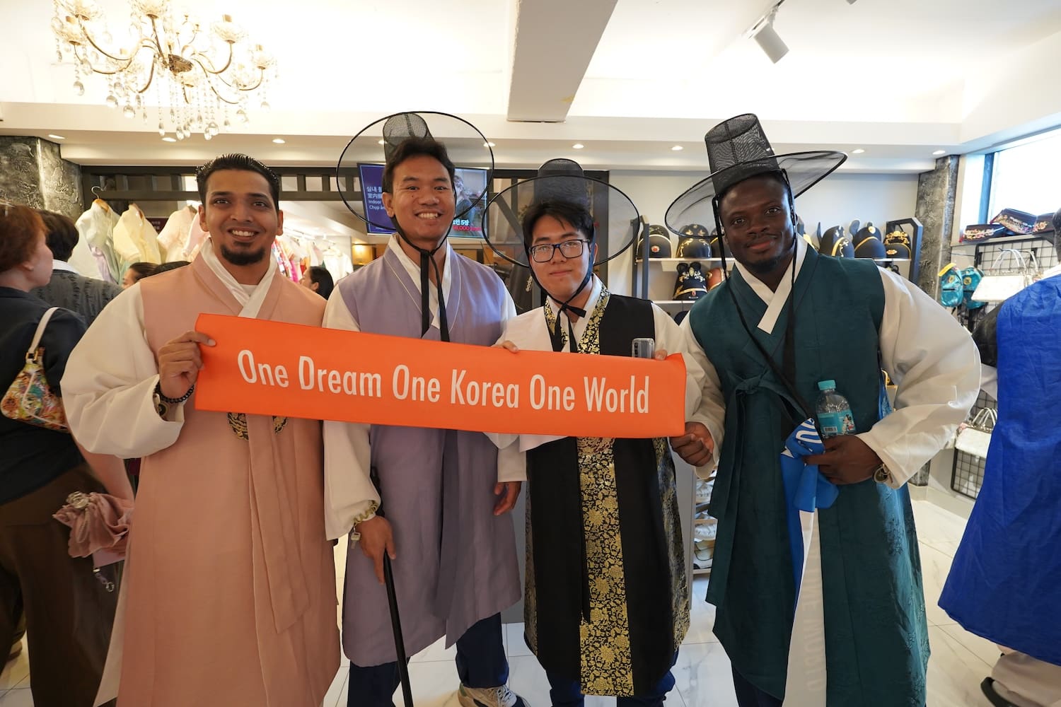 Four men in traditional Korean hanbok hold an orange banner reading “One Dream One Korea One World” inside a shop, supporting Action for Korea United and the global push for Korean reunification.