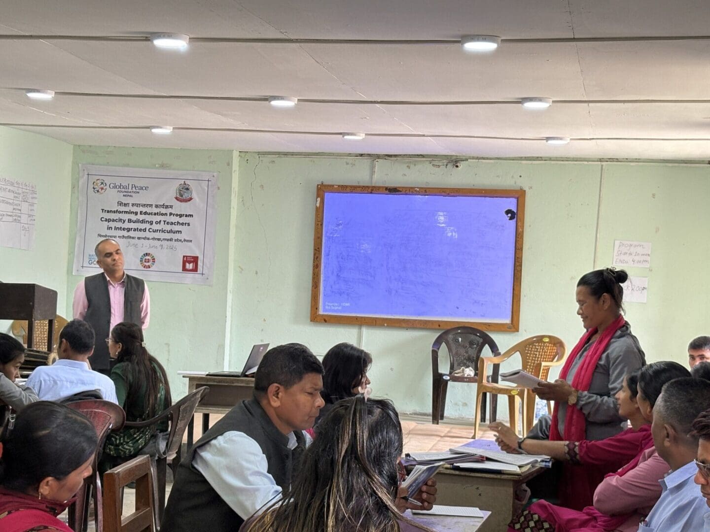 A group of adults attend a capacity building training session in a classroom in Nepal; a woman stands reading a paper while a man stands near a sign and a digital screen.